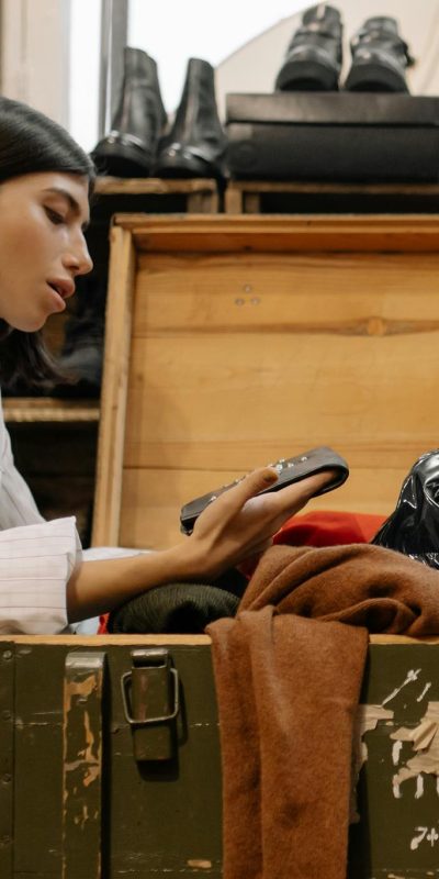 A woman examines clothes from an open chest in a thrift store filled with shoes.