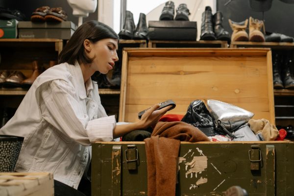 A woman examines clothes from an open chest in a thrift store filled with shoes.